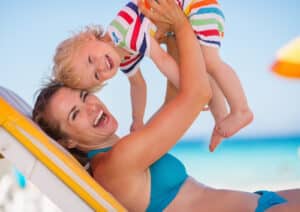 Woman holds baby while sitting on a chair at the beach in a bikini after tummy tuck.